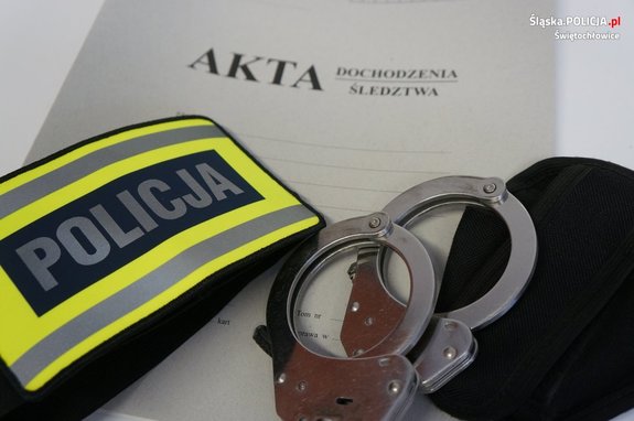 zdjęcie przedstawia: kajdanki i opaska odblaskowa z napisem policja, leżące na aktach dochodzeniowo-śledczych