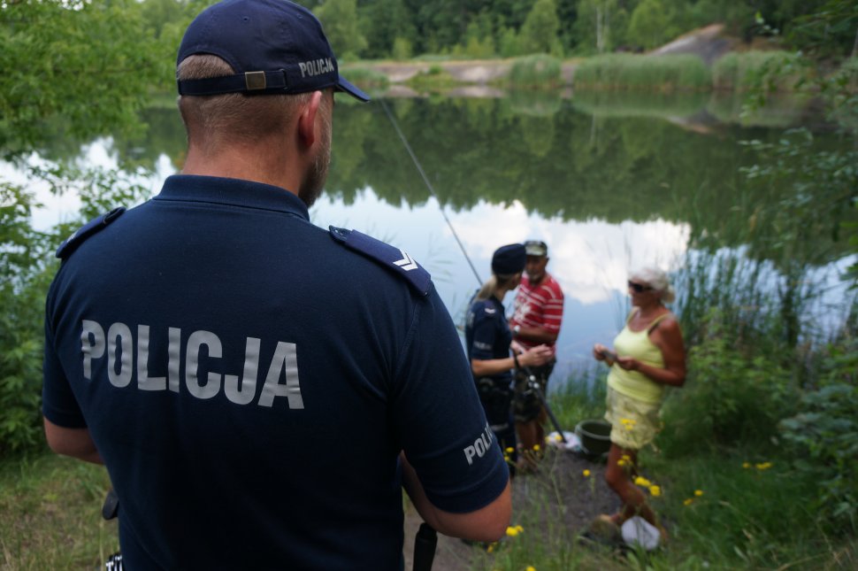 Na pierwszym planie zdjęcia umundurowowany policjant obserwujący sytuację nad akwenem. W tle widac policjantkę która profilaktycznie przekazuje bilet na basen osobą odpoczywającym w rejonie zbiornika wodnego 