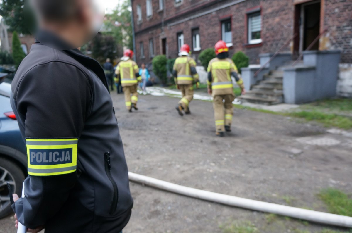 Na pierwszym planie zdjęcia widać nieumundurowanego policjanta z opaską identyfikacyjną na ramieniu z napisem policja, w tle widać pozostałe służby pracujące przed jedynym z budynków w Świętochłowicach