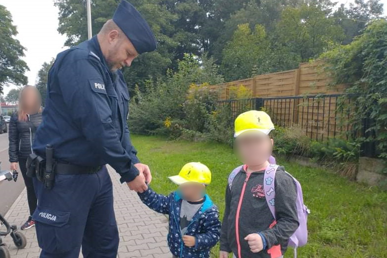 Na zdjęciu mundurowy zakłada na ręce chłopcu silikonową opaskę z kampanii Mamo! Tato! Tutaj jestem ! Chłopiec stoi obok dziewczynki, oboje mają na głowie czapki odblaskowe z logo kampanii. W tle widać mamę dzieci. 