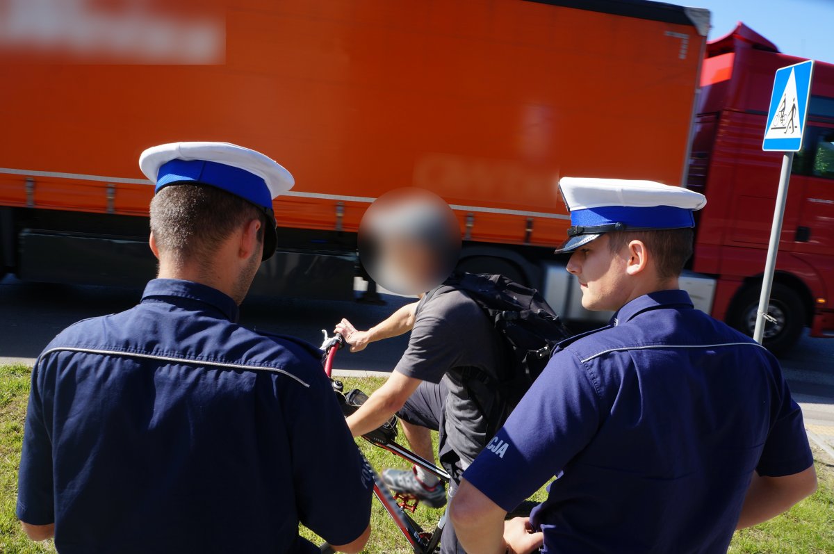 Na zdjęciu widac jak policjanci kontrolują w trakcie działań rowerzytę. W tle widac przejeźdzający pojazd ciężarowy 