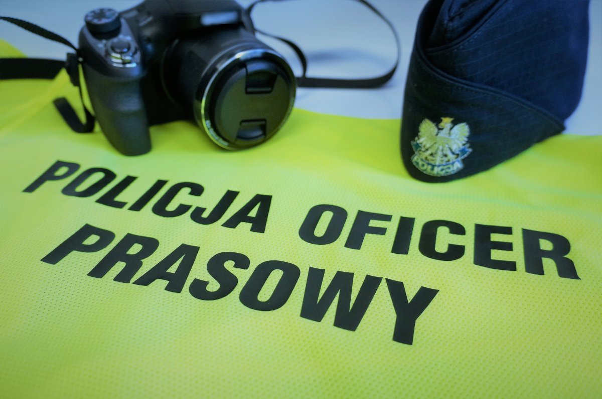 Na zdjęciu kamizelka odblaskowa z napisem POLICJA OFICER PRASOWY,  w tle z lewej strony aparat fotograficzny, z prawej czapka służbowa koloru granatowego z orzełkiem