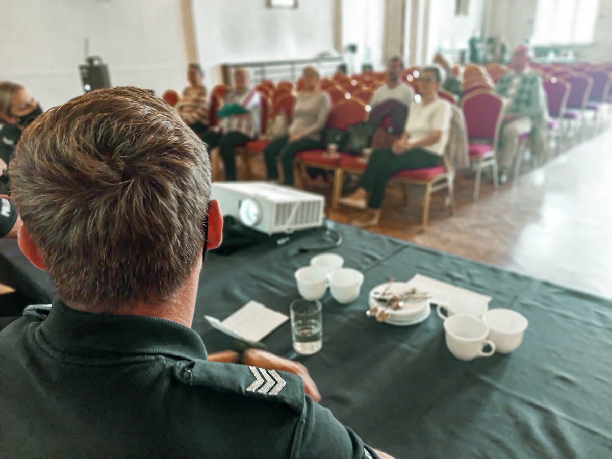 Na zdjęciu po lewej stronie na pierwszym planie mundurowy siedzący tyłem do obiektywu, lecz twarzą do słuchaczy. W tle widac siedzacych seniorów, przysłuchujących się prowadzonemu wykładowi oraz umundurowaną policjantkę 