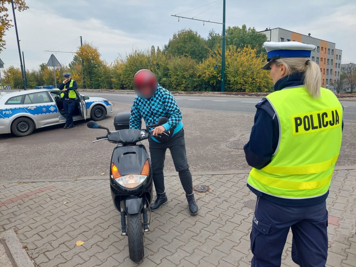 Na zdjeciu policjantka w trakcie konbtroli dorogowej kierującego motorowerem. W tle widać radiowóz policyjny oraz mundurowego, który podaje informacje dyżurnemu