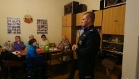 Dzielnicowy stojący bokiem, w tle seniorzy siedzący przy stole.