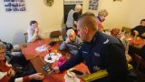 Na zdjęciu dzielnicowy rozdający odblaski oraz ulotki, w tle siedzący przy stole seniorzy z Polskiego Towarzystwa Osób Niewidomych i Niedowidzących.