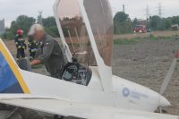 pilot oraz samolot dwumiejscowy po awaryjnym lądowaniu na nieużytkach, w tle strażacy