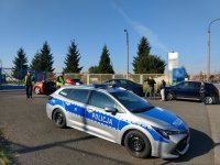 Policjanci z Wydziału Ruchu Drogowego KMP Świętochłowice podczas wykonywania czynności służbowych po zatrzymaniu pojazdu do kontroli. W tle zaparkowany kontrolowany pojazd oraz ekipa TVP1 w trakcie nagrania.