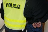 zatrzymany mężczyzna zakuty w kajdanki z rękoma w tyle w asyście policjanta z wydziału kryminalnego, ubranego w kamizelkę odblaskową z napisem policja