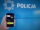 Na zdjęciu widać dłoń trzymająca telefon komórkowy, w tle napisz POLICJA