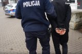 Na zdjęciu policjant podczas doprowadzania zatrzymanego mężczyzny do jednostki policji. Zatrzymany ma założone kajdankami na rękach trzymanych z tyłu.