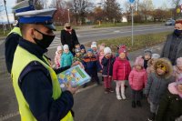 Na zdjęciu policjanci wraz z grupą przedszkolaków oraz opiekunów po prelekcji na temat bezpiecznego przechodzenia przez jezdnię. Na zdjęciu jedne z policjantów trzyma w rękach kilka elementarzy bezpieczeństwa
