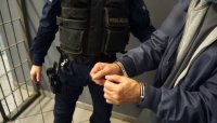 zatrzymany mężczyzna zakuty w kajdanki w asyście umundurowanego policjanta na tle policyjnej celi