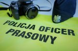 Na zdjęciu kamizelka odblaskowa z napisem policja oficer prasowy. Na drugim planie po prawej stronie czapka - furażerka z orłem na przodzie, po lewej stronie aparat