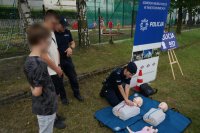 Na zdjęciu mundurowa prowadzi pokaz pierwszej pomocy, w tym resuscytacji krążeniowo oddechowej na fantomie, w tle widać innego policjanta oraz uczestników festynu przyglądających się czynnością wykonywanym przez policjantkę