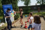 Na zdjęciu mundurowa, która stroi przy kole wiedzy z listą pytań, obok stoją dzieci czekające w kolejnce do zakręcenia kołem aby odpowiedzieć na pytania związane z bezpieczeństwem