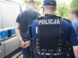 Na zdjęciu umundurowany policjant, przed nim nieumundurowany policjant asekuruje osobę zatrzymaną zakutą w kajdanki. Po lewej stronie widać fragment radiowozu