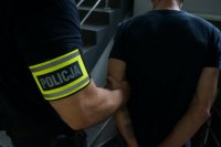 Na zdjęciu nieumundurowany policjant z żółta opaską na ramieniiu z napisem policja prowadzi zatrzymanego mężczyznę zakutego w kajdanki na ręce trzymane z tyłu