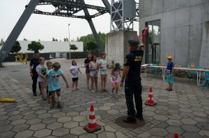 Na zdjęciu dzieci biorące udział w sztafecie z goglami na twarzy które imitują stan po spożyciu używek. Strażnik miejski trzyma uniesioną rekę ku górze w której trzyma różową wstążkę i czeka na przygotowanie dzieci aby rozpocząć rywalizację