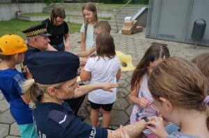 Zdjęcie przedstawia dzieci uczestniczące w zajęciach wakacyjnych, które naklejają mundurowym na rękach konfetti które jest elementem zabawy polegającej na tym że wygrywa ta drużyna która przyklei więcej konfetii.