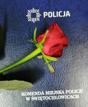 Na zdjęciu część teczki koloru granatowego z napisem Policja i znakiem graficznym gwiazdy policyjnej, tak zwana gwiazda ośmioramienna, na teczce czerwona róża