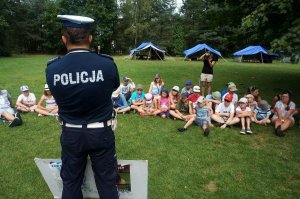 Na zdjęciu policjant na spotkaniu profilaktycznym dotyczącym bezpiecznych wakacji. W tle młodzeż siedzaca na trawie, uczestnicząca w spotkaniu