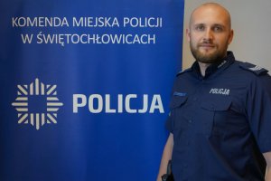 sierż. szt. Rafał Kamiński na tle niebieskiego banera z napisem Policja i Komenda Miejska Policji w Świętochłowicach