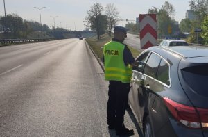 Na zdjęciu mundurowy w trakcie kontroli drogowej zatrzymanego pojazdu. Po prawej stronie widac drogę o 3 pasach ruchu w jednym kierunku