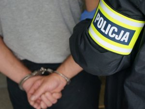 Na zdjęciu zbliżenie na ręce zatrzymanego mężczyzny, zakutego w kajdanki, który prowadzony jest przez nieumundurowanego policjanta z opaską odblaskową z napisem policja na ramieniu