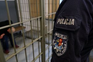 Na zdjęciu ramie umundurowanego policjanta na którym widać napis policja oraz naszywkę identyfikującą Komendę Miejską Policji w Świętochłowicach z gwiazdą policyjną i herbem miasta.  W tle widać zatrzymanego mężczyznę, który przebywa w pomieszczeniu dla osób zatrzymanych