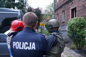 Na zdjęciu widac stojących przez budynkiem mundurowych tj. strażak, miejscowy policjant oraz funkcjonariusz Samodzielnego Pododdziału Kontrterorystycznego Policji w trakcie uzgadniania taktyki działania podczas prowadzonej interwencji