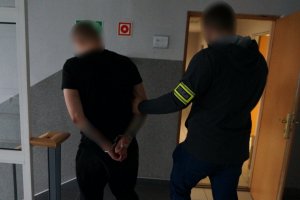 Na zdjęciu nieumundurowany policjant z opaską odbalskową na ramieniu podczas prowadzenia zatrzymanego meżczyzny zakutego w kajdanki na ręce trzymane z tyłu.