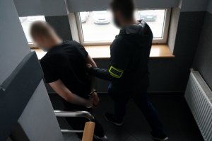 Na zdjęciu nieumundurowany policjant z opaską odbalskową na ramieniu podczas prowadzenia zatrzymanego meżczyzny zakutego w kajdanki na ręce trzymane z tyłu.