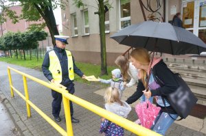 Na zdjęciu policjant oraz dwie kobiety z dziećmi. Mundurowy przekazuje dzieciom gadżety profilaktyczne w ramach akcji bezpieczna droga do szkoły