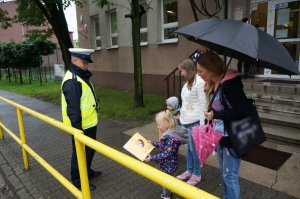 Na zdjęciu policjant oraz dwie kobiety z dziećmi. Mundurowy przekazuje dzieciom gadżety profilaktyczne w ramach akcji bezpieczna droga do szkoły