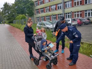 Mundurowy w trakcie rozmowy z rodzicami w dniu rozpoczęcia roku szkolenego w jednej ze świętochłowickich szkół. Na zdjęciu widać jak mundurowy przekazuje dzieciom gadżety związane z bezpieczeństwem