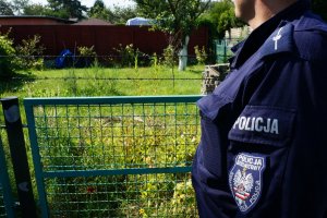 Zbliżenie na ramie umundurowanego policjanta. Na zdjęciu widać pagon ze stopniem młodszego aspiranta, poniżej kieszeń na ramieniu z napisem policja, pod naszywka z napisem Dzielnicowy oraz Komenda Miejsa Policji. W tel widać ogródek działkowy