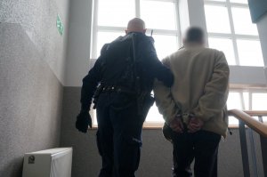 Na zdjęciu umundurowany policjant prowadzi zatrzymanego mężczyznę zakutego w kajdanki na ręce trzytmane z tyłu