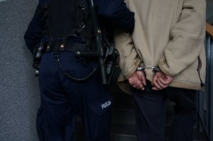 Na zdjęciu zbliżenie na umundurowanego policjanta, ktory prowadzi zatrzymanego mężczyznę zakutego w kajdanki na ręce trzytmane z tyłu