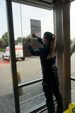 Na zdjęciu mundurowa wiesza plakat kampanii „Mamo! Tato! Tutaj jestem!” na drzwiach centrum handlowego