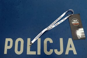 Na zdjęciu na granatowym tle napis Policja, w órnym prawym rogu rozłożona smycz do kluczy z napisem „Pasy. To trzyma cię przy życiu” oraz czarna ulotka z rysunkiem pojazdu i napisem Twoje światła, twoje bezpieczeństwo.