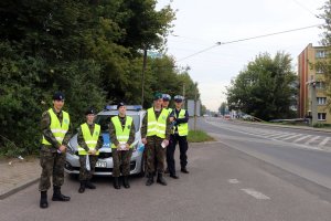 Na zdjęciu ulica, z lewej strony na poboczu stoi oznakowany radiowóz, przed nim czwórka uczniów w mundurach szkolnych moro i kamizelkach odblaskowych, za nimi dwóch umundurowanych policjantó w kamizelkach odblaskowych