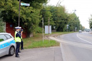 Na zdjęciu ulica, na poboczu z lewej strony stoi oznakowany radiowóz, przy nim umundurowany policjant w kamizelce odblaskowej, który trzyma w ręce urządzenie do mierzenia prędkości