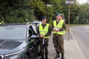 Na zdjęciu ulica, na poboczu z lewej strony na pierwszym planie dwóch uczniów w mundurach i kamizelkach odblaskowych, jeden z nich trzyma w lewej ręce smycze do kluczy, drugi trzyma ulotki.Stoją obok zatrzymanego do kontroli pojazdu. Na drugim planie w tle widać policjanta ruchu drogowego w czapce służbowej, część radiowozu z napisem policja oraz jednego ucznia w czapce ze szkolnego umundurowania.