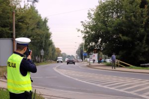 Na zdjęciu ulica, na lewym poboczu stoi umundurowany policjant w kamizelce odblaskowej z napisem Policja, trzyma w ręce urządzenie do mierzenia prędkości-skierowane w stronę porusząjących się pojazdów