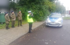 Na zdjęciu ulica, na lewym poboczu oznakowany radiowóz marki toyota, przed nim stoi umundorowany policjant w kamizelce odblaskowej z napisem Policja, który trzyma w ręce urządzenie do mierzenia prędkości skierowane na nadjeżdżajace pojazdy. Z lewej strony radiowozu stoi trzech uczniów w mundurach szkolnych moro i kamizelkach odblaskowych