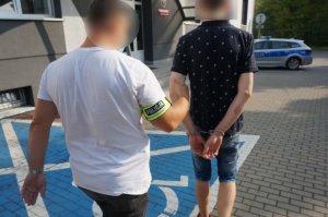 Na zdjęciu parking przed Komendą Policji w Świętochłowicach. W kierunku wejścia kieruje się nieumundurowany policjant w białej koszulce z krótkim rękawem i dżinsach, na prawym ramieniu ma opaskę odblaskową z napisem Policja.Prowadzi zatrzymanego mężczyznę. Zatrzymany znajduje się z prawej strony policjanta, widoczny jest od tyłu,ma kajdanki założone na ręce trzymane z tyłu. Ubrany jest w granatową koszulkę z krótkim rękawem w białe wzory i dżinsowe spodenki do kolan oraz sportowe buty.