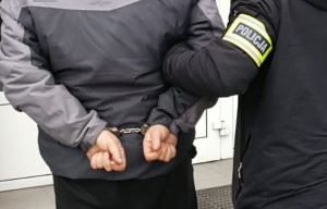 Na zdjęciu zbliżenie na ręce zatrzymanego mężczyzny, prowadzonego przez nieumundurowanego policjanta. Policjant ma założoną opaskę odblaskową z napisem Policja na ramieniu