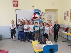 Na zdjeciu wnętrze jednej z klas. Dzieci stoja wraz z maskotka śląskiej Policji w grupie do pamiątkowego zdjęcia. Uczestnicy zajęć trzymają w dłoniach elementarze bezpieczeństwa które otrzymały od mundurowych. Jedna z dziewczynek trzyma w rękach miniaturową maskotkę, odzwierciedlającą Psa Sznupka