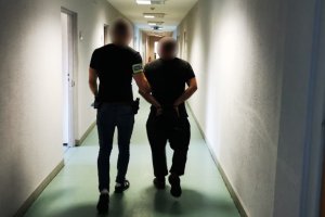 Na zdjęciu neumundurowany policjant z opaska odblaskową z napisem policja na prawym ramieniu prowadzi po korytarzu zatrzymanego mężczyznę zakutego w kajdanki na ręce trzemane z tyłu